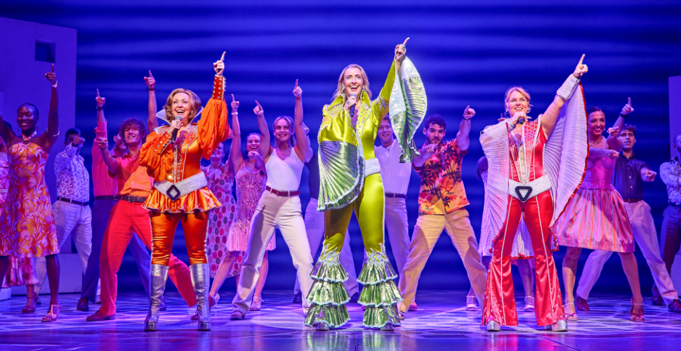 A group of performers on stage in vibrant, 1970s-inspired costumes. Three lead performers in orange, green, and red outfits with flared pants and platform boots stand at the front, each raising one arm with an index finger pointing upwards. Behind them, more performers in colorful attire strike similar poses against a blue gradient backdrop, creating a lively and energetic scene